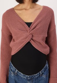 Gestrickte, mauvefarbene, verkürzte Strickjacke mit einem verdrehten Vorderdesign und langen Ärmeln, getragen über einem schwarzen Oberteil und blauen Jeans. Strukturierten Stoff.