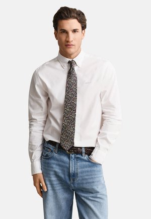Homme portant une chemise blanche à boutons, une cravate à motif paisley, une ceinture tressée marron et un jean bleu clair sur fond blanc.