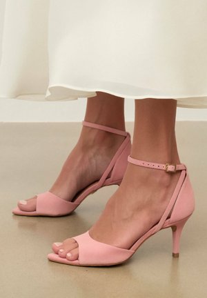 Stopy w różowych zamszowych sandałach na obcasie z odkrytymi palcami i paskami wokół kostki, stojące na gładkiej podłodze pod białą sukienką.