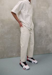 Personne portant une chemise à manches courtes écrue et un pantalon avec des chaussures de sport blanches, noires et rouges, debout contre un mur gris texturé.