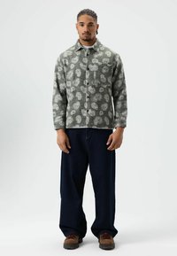 Grau gemusterte Hemdjacke mit hellen Spiraldesigns, Kragen, einzelne Brusttasche, kombiniert mit dunklen, weit geschnittenen Jeans und braunen Schuhen.