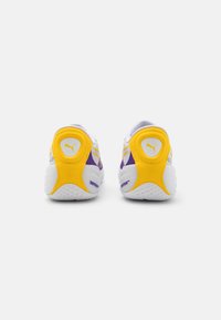 Adidași Puma cu un superior predominant alb, cu accente galbene și violete, talpă din cauciuc texturat și un logo circular pe călcâi.