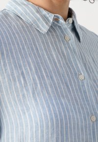 Gros plan d'une personne portant une chemise bleu clair à boutons avec des rayures blanches verticales et un col classique.