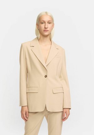 Beige skräddarsydd blazer med en ensam brun knapp, breda slag och två framfickor; slät struktur och en välarbetad passform.