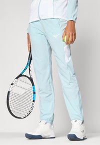 Lysblå træningsbukser med hvide detaljer og en lynlås lomme, parret med hvide sportssko og et tennisracket, mens der holdes en tennisbold.