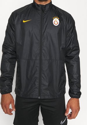 Homme portant une veste de sport noire zippée avec le logo jaune Nike et le logo du club de football Galatasaray sur la poitrine.