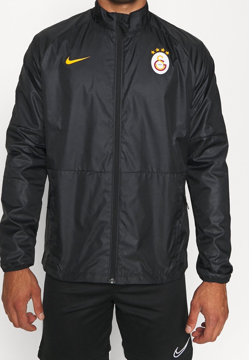 Homme portant une veste de sport noire zippée avec le logo jaune Nike et le logo du club de football Galatasaray sur la poitrine.
