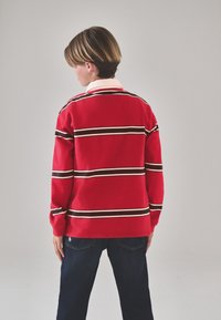 Camiseta roja de manga larga con rayas horizontales blancas y marrón oscuro, con cuello con solapa. Combinada con vaqueros de mezclilla oscuros.