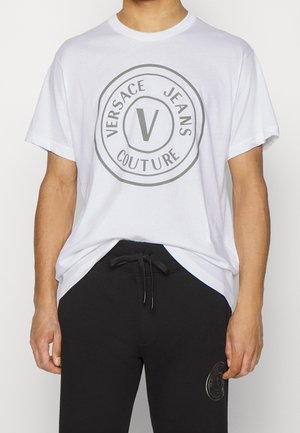 Weißes Baumwoll-T-Shirt mit einem runden grauen "Versace Jeans Couture"-Logo auf der Brust, kombiniert mit schwarzen Jogginghosen mit einem Logodetail.