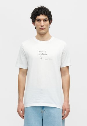 Άνδρας με σγουρά μαλλιά που φοράει λευκό μπλουζάκι με την επιγραφή «Limited Edition» και ανοιχτά μπλε τζιν, στέκεται μπροστά σε απλό λευκό φόντο.