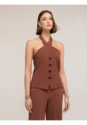 Kvinde iført brun ærmeløs haltertop med knapper og matchende bukser, accessoriseret med guldbelagte øreringe og armbånd, foran en ensfarvet baggrund.