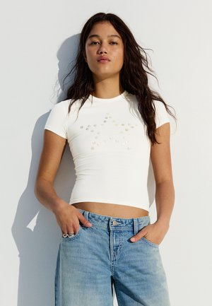 Fiatal nő hosszú sötét hajjal, fehér, csillagmintás rövid felsőt és világoskék farmert visel, egy fehér falnak támaszkodik.