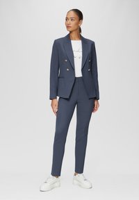 Blazer ajusté bleu marine avec des boutons dorés, associé à une chemise graphique blanche et un pantalon slim, complété par des baskets blanches imposantes.