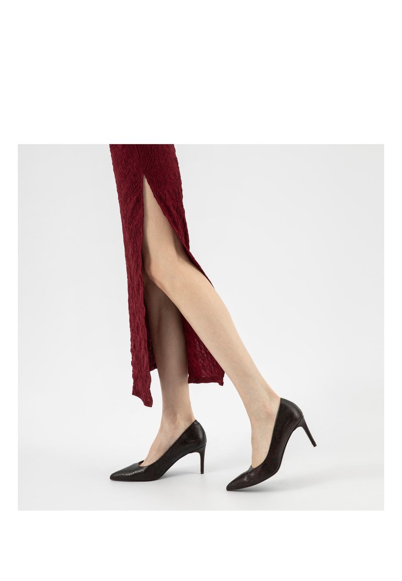 Vrouw met zwarte pumps met een puntige neus, gecombineerd met een donkerrode rok met structuur en een hoge, zijsplit.