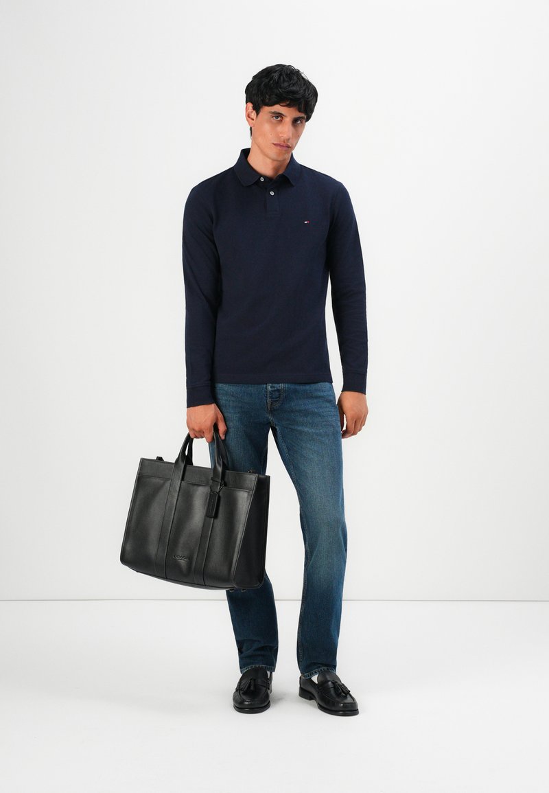 Man med marinblå långärmad polo, blå jeans, svarta loafers, och håller en svart läderhandväska framför en enfärgad vit bakgrund.