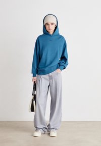 Felpa blu con cappuccio e logo, jeans grigi a vestibilità ampia, berretto in maglia crema e scarpe bianche. Tiene in mano una borsa nera con accenti crema.