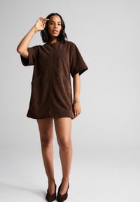 Robe mini en daim marron avec des manches courtes, présente une coupe ample et deux poches avant. Texture lisse avec des détails de couture minimaux.