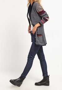 Dzianinowy kardigan w granatowym kolorze z wzorzystymi rękawami, kieszeniami i kołnierzem typu szal. Połączony z ciemnymi dżinsami i czarnymi botkami za kostkę.