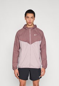 Kerge kapuutsiga jakk mauve ja heleroosa toonides, millel on esil tõmblukk, Nike logo rinnal ja elastsed mansetid mugavaks sobivuseks.