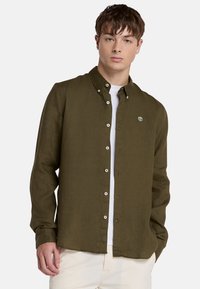 Camisa de color verde olivo, con botones, cuello clásico, mangas largas y un pequeño parche con el logo en el pecho. Tejido ligero y texturizado.