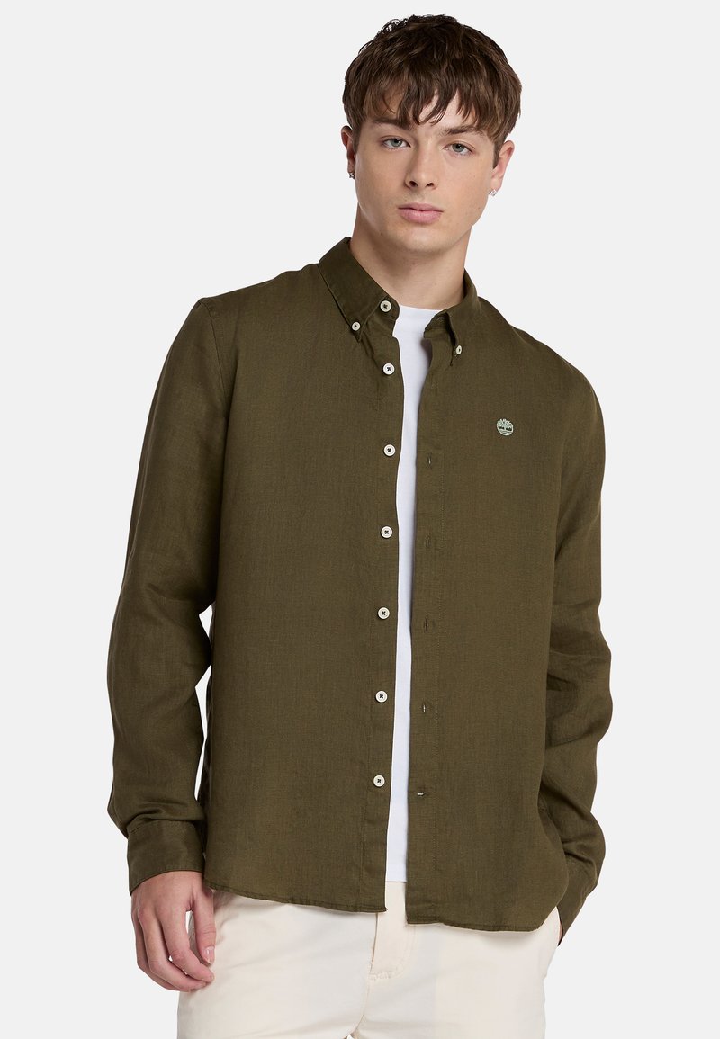 Camisa de color verde olivo, con botones, cuello clásico, mangas largas y un pequeño parche con el logo en el pecho. Tejido ligero y texturizado.