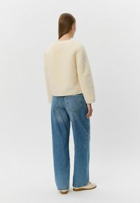 Krémovo sfarbený, textúrovaný fleece sveter s dlhými rukávmi, v kombinácii s voľnými modrými džínami a béžovými topánkami, z pohľadu zboku.