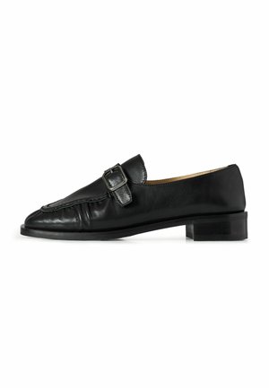 Mocasín de cuero negro con tacón bajo y detalle de hebilla en la correa superior, mostrado de lado sobre un fondo blanco.