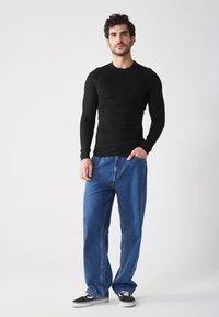 Haut noir côtelé à manches longues associé à un jean bleu ample en denim et des baskets noires, posé devant un fond clair neutre.