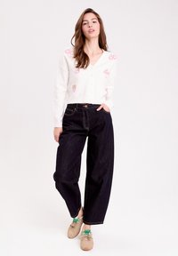 Gilet en tricot blanc avec broderies florales, associé à un pantalon large en denim foncé. Le modèle porte des chaussures beige avec des lacets verts.