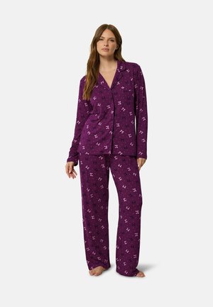 Femme portant un pyjama violet à manches longues boutonné et un pantalon assorti avec de petits imprimés papillons, debout pieds nus sur un fond uni.