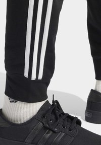 Svarta sportskor med tre mörkgrå randdetaljer, i kombination med svarta joggers med vita ränder och ljusa randiga strumpor.