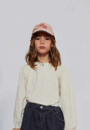 Top de manga larga en beige claro con hombros fruncidos, combinado con una falda vaquera oscura. La modelo lleva una gorra rosa claro con un logo de texto rojo.