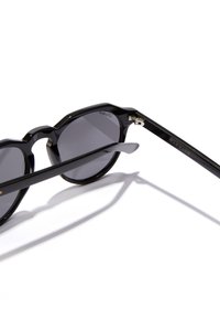 Mustat aurinkolasit pyöreillä linsseillä, paksuilla kiiltävillä kehyksillä ja huomaamattomalla kultaisella logolla varressa. Linssit ovat tummanharmaat, tarjoavat UV-suojan.