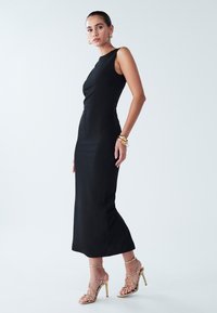 Black, form-fitting sleeveless dress with a high neckline, smooth texture, and a subtle mermaid hem, paired with gold strappy high heels.