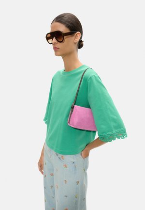 Kobieta w dużych brązowych okularach przeciwsłonecznych, zielonej bluzce z krótkim rękawem i koronkowymi wykończeniami, jasnoniebieskich spodniach w kwiaty oraz trzymająca różową torbę na ramię.