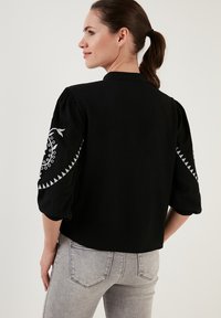 Schwarze, kurz geschnittene Jacke mit puffigen Ärmeln, die weiße Stickerei in einem kreisförmigen Muster auf den Ärmeln aufweist. Strukturierter Stoff, strukturiertes Design.