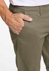Pantaloni verde oliva con finitura testurizzata, dotati di tasche laterali e vita aderente. Mano visibile appoggiata nella tasca.