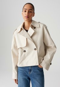 Cappotto trench corto beige con ampio colletto, bottoni doppi e spalline. Indossato sopra una maglia scura e jeans blue.