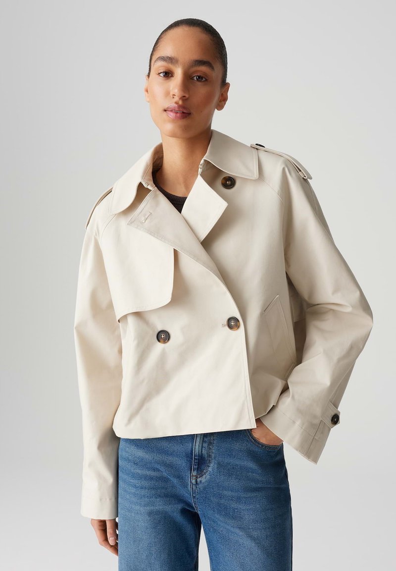 Cappotto trench corto beige con ampio colletto, bottoni doppi e spalline. Indossato sopra una maglia scura e jeans blue.