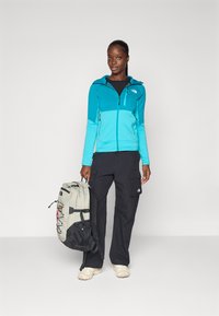 Felpa con zip turchese, pantaloni cargo neri, in mano uno zaino grigio con dettagli. Tessuto liscio, cappuccio regolabile, due tasche laterali con zip.