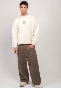 Kremowa bluza z haftowanym złotym logo, zestawiona z luźnymi brązowymi spodniami typu cargo z dużymi bocznymi kieszeniami i swobodnym krojem.