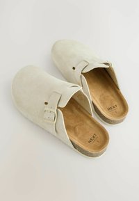 Sabots en suède beige clair avec une sangle à boucle, bout rond et semelle intérieure rembourrée marron. Semelle texturée pour une bonne adhérence. Nom de la marque imprimé à l'intérieur.