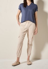 Lehká modrá košile s límečkem, volný střih; v kombinaci se béžovými elastickými cargo kalhotami s kapsami a zúženými lemy. Hnědé slip-on boty.