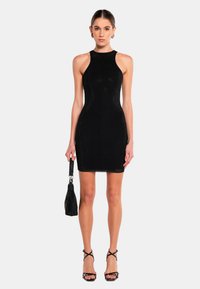 Robe noire à dos nu avec une coupe ajustée, un tissu texturé et un léger éclat. Associée à des sandales noires à lanières et à un petit sac.