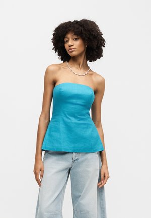 Γυναίκα με σγουρά μαλλιά που φοράει ένα τιρκουάζ τοπ χωρίς τιράντες και ανοιχτόχρωμα τζιν, στέκεται μπροστά σε ένα απλό λευκό φόντο.