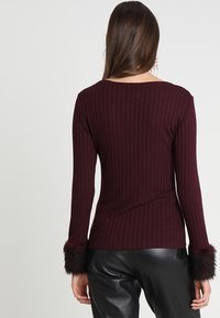 Vrouw met lang bruin haar draagt een aansluitende, geribbelde donkerbordeaux trui met bontmanchetten en zwarte leren broek, met de rug naar de camera.