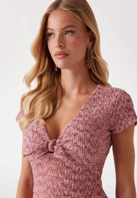 Rose-colored dress with a textured fabric, short sleeves, and a v-neckline featuring a knotted detail at the chest.