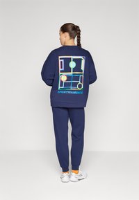 Marineblå hettegenser og bukser, med et fargerikt geometrisk design på baksiden. Hvite sneakers med oransje detaljer fullfører antrekket.