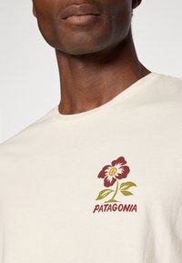 Krēmos krāsas kokvilnas T-krekls ar ziedu grafiku bordo un zaļā krāsā. Zem dizaina atrodams teksts "PATAGONIA".