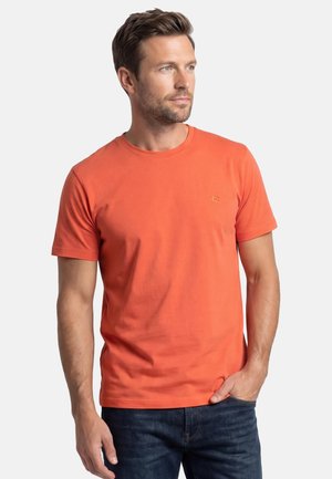 Mann mit kurzem braunem Haar, der ein oranges kurzärmeliges T-Shirt und dunkle Jeans trägt, steht mit einer Hand in der Tasche und schaut nach rechts.